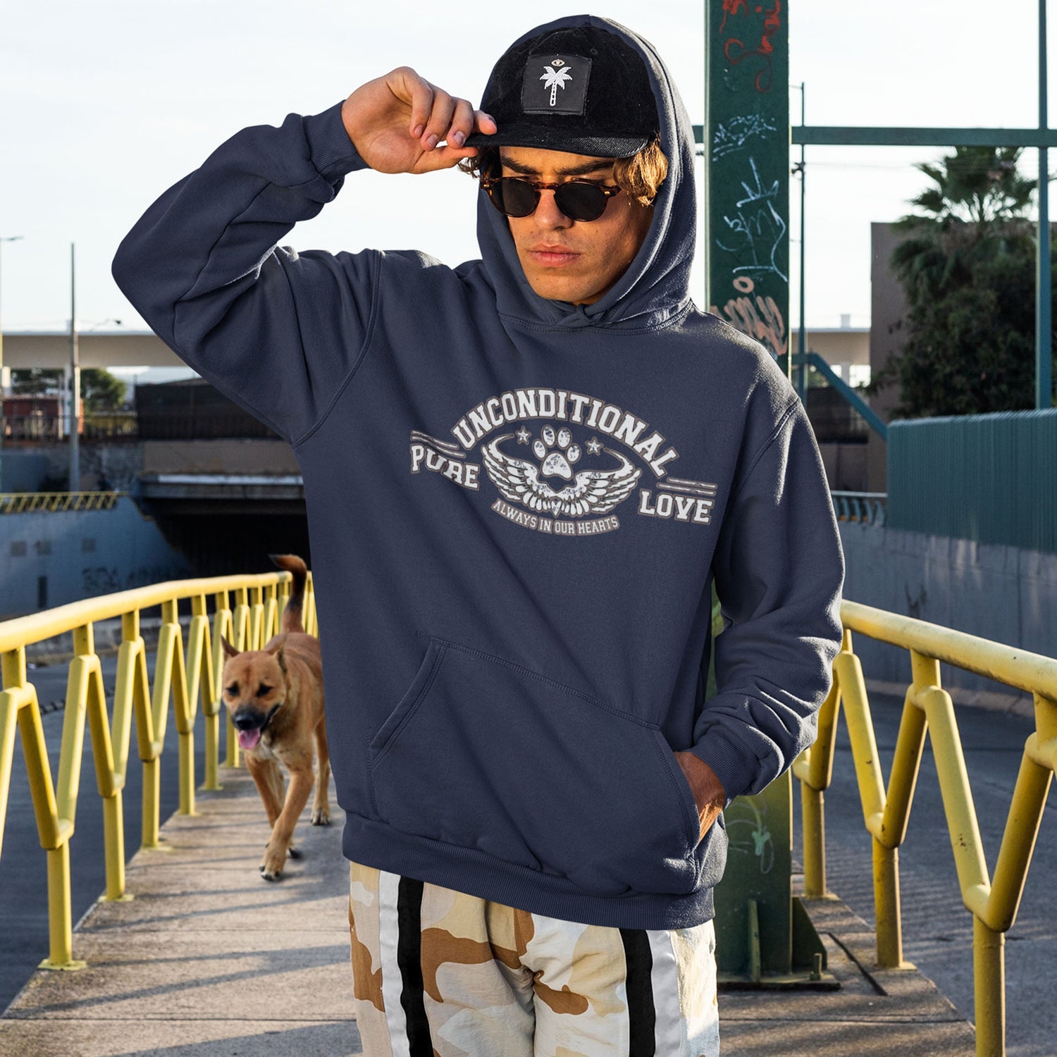  A young man strolls across a bridge, one hand reaching up to adjust his cap, while his faithful dog follows closely behind. He sports sunglasses and a stylish navy Dogs Pure Love hoodie.