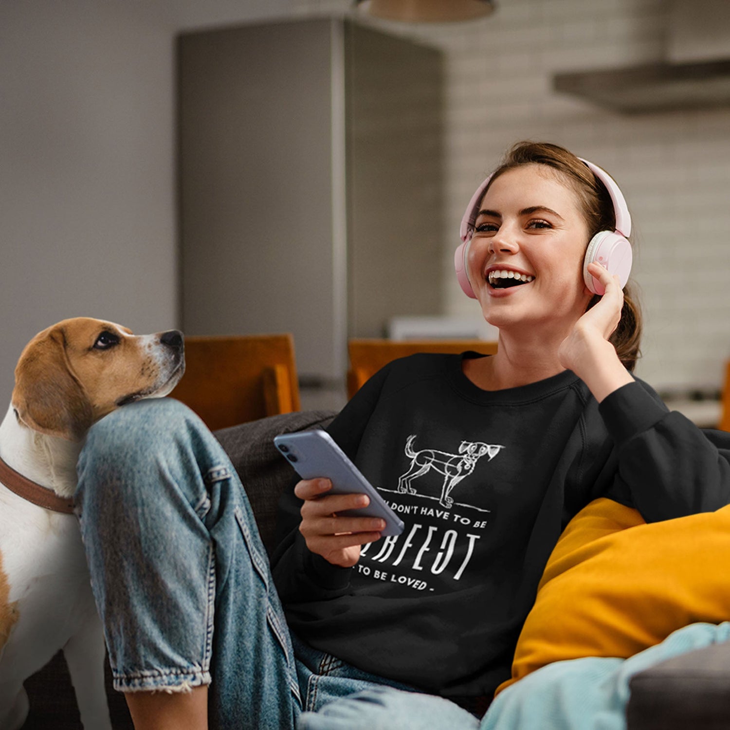 A young woman wearing headphones listens to her music smiling as she holds her phone. Beside her, her Beagle companion sits contentedly on the couch, resting its head against her uplifted knee.
