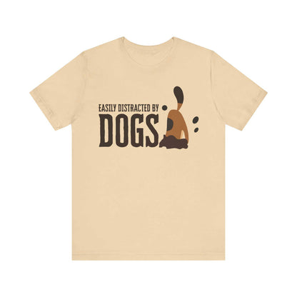 Amidst a white setting, a Dogs Pure Love soft cream unisex t-shirt features a fun slogan ‘Easily Distracted by Dogs,’ and a graphic of a dog digging.