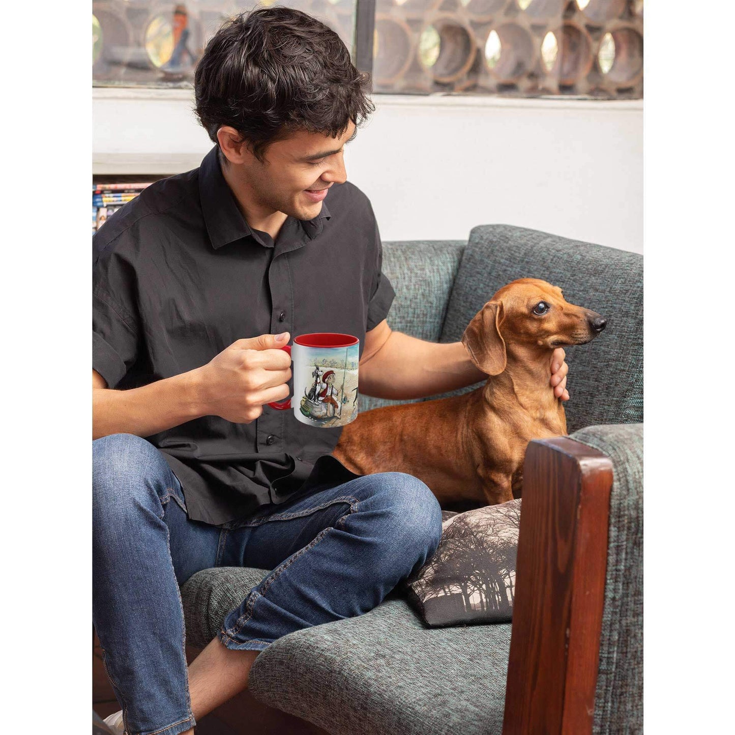 A man sits, cradling his Dogs Pure Love ceramic mug adorned with an illustration, while he glances to his side, gently patting his Dachshund resting on a lounge chair.