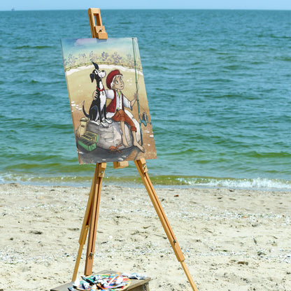 'Dogs Pure Love' canvas of a boy and dog fishing displayed on a wooden artist easel is showcased on a sandy beach.