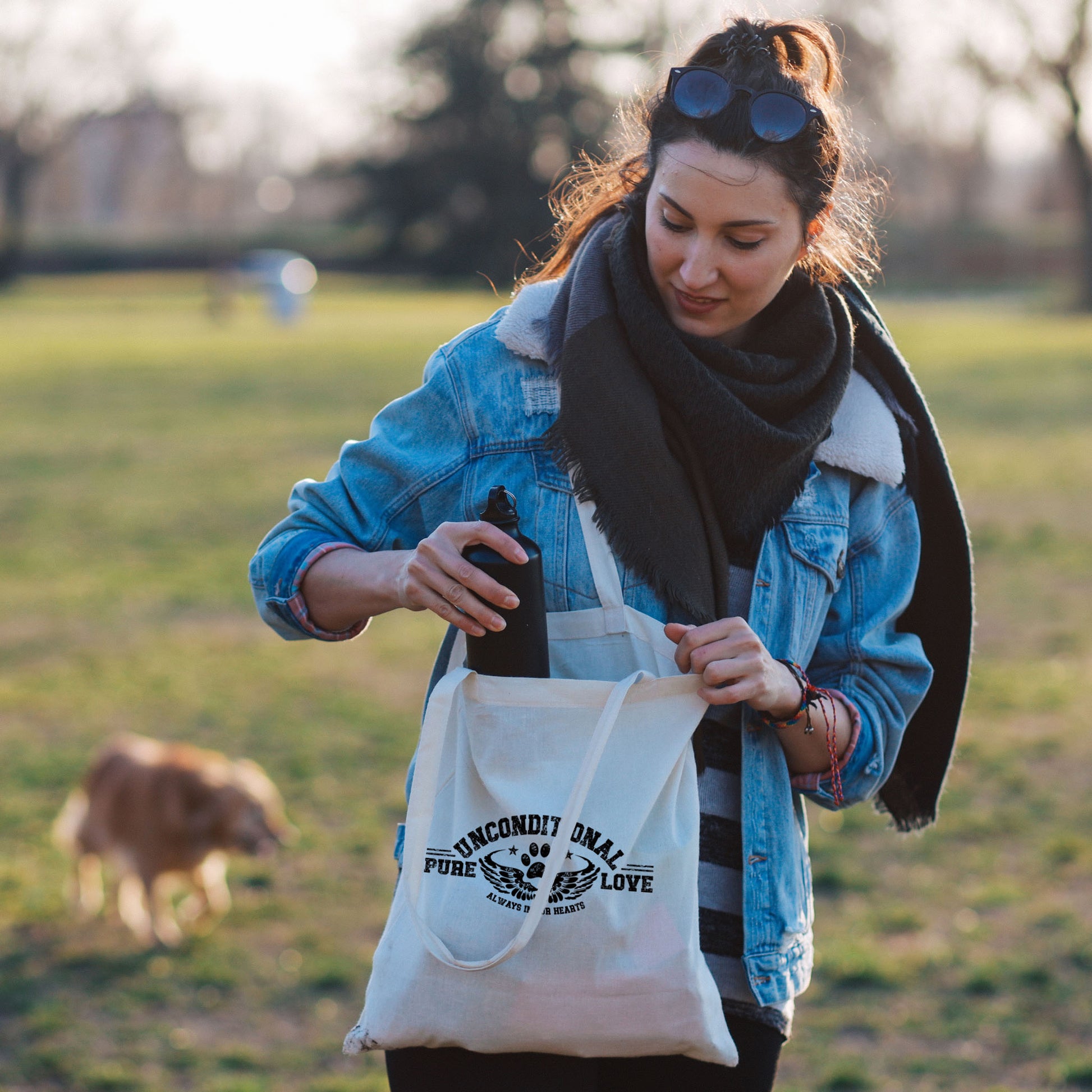 A woman strolls with her dog in the park, pausing to peer into her Dogs Pure Love tote bag. With a gentle gaze downward, she retrieves her water bottle.