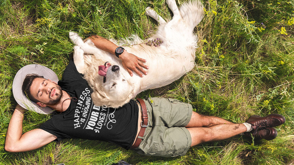 A man lays on the grass wearing a black Dogs Pure Love tee with one arm behind his head, while the other embraces his Golden Retriever.