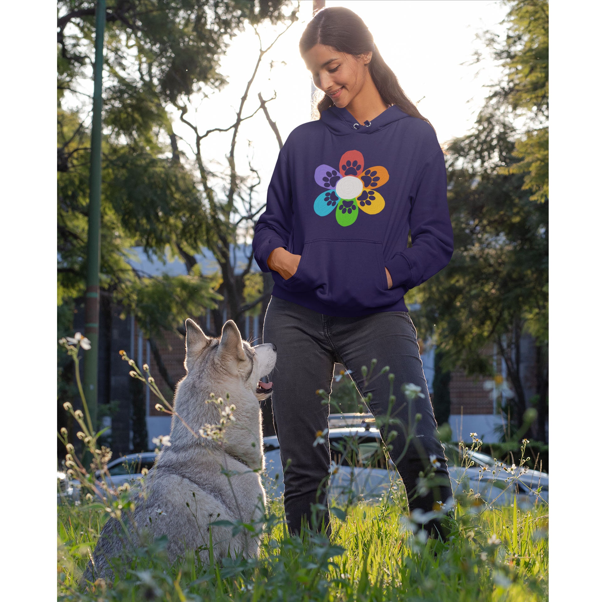 A young woman, dressed in a Dogs Pure Love flower paw print hoodie, stands in a garden with her hands in her pockets. She looks down at her dog, who gazes up at her with adoration.