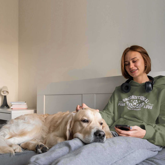 A woman lays on top of a bed patting her Golden Retriever as it lays its head on her lap. The woman's headphones lay around her neck as she smiles at her dog. She is wearing a Dogs Pure Love military green custom dog hoodie with the words 