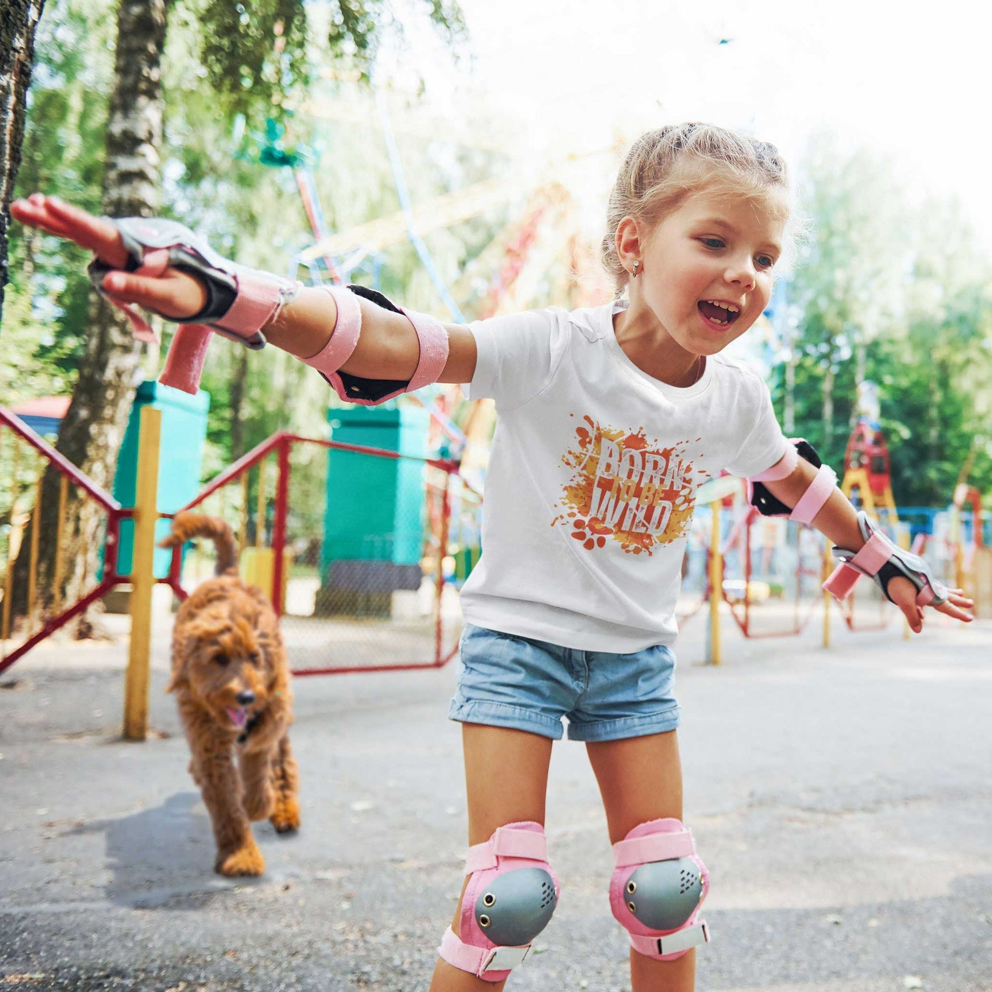  A young girl, dressed in a Dogs Pure Love white tee featuring the 'Born to be Wild' print, roller skates with her arms outstretched at a park, with her dog following behind her.