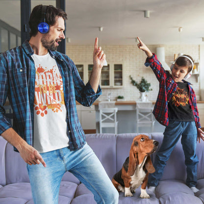  In a lounge, a father dances while his son dances on the couch, both pointing upwards, wearing headphones and Dogs Pure Love tees with the 'Born to be Wild' print. Their Basset Hound sits on the couch, gazing up at the boy.