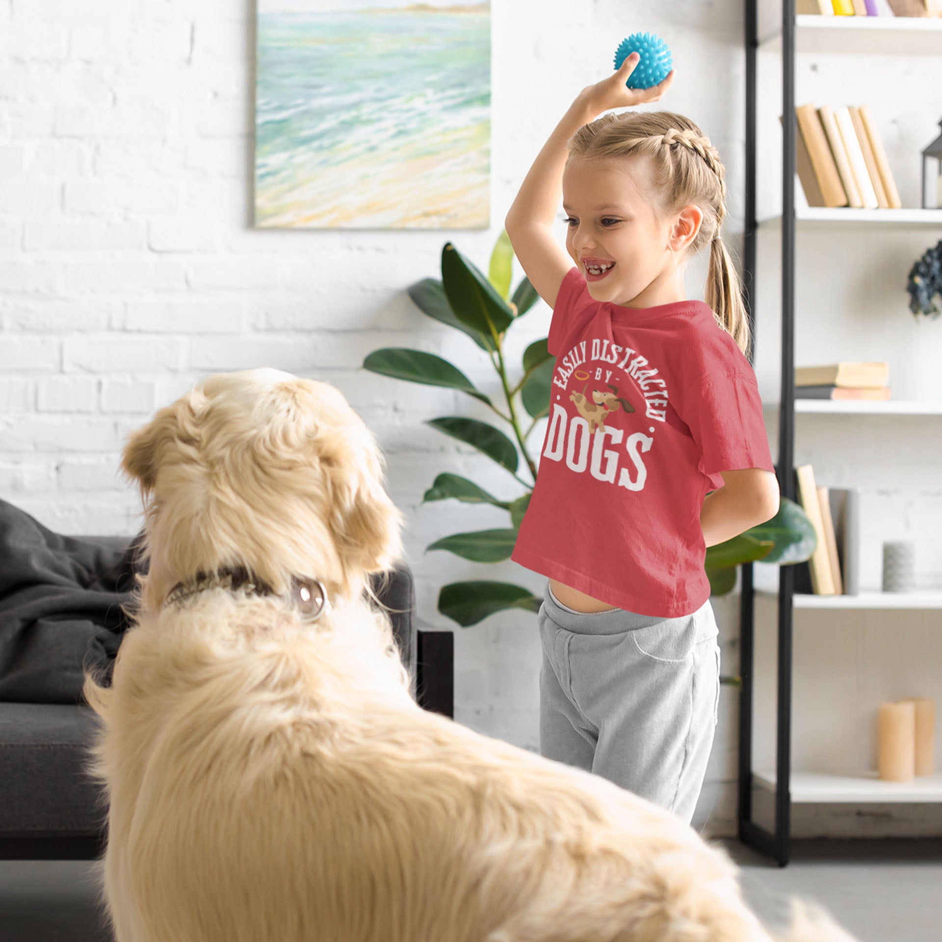  A young girl, clad in a Dogs Pure Love tee with the print 'Easily Distracted by Dogs,' holds up a ball in the air in front of a Golden Retriever.
