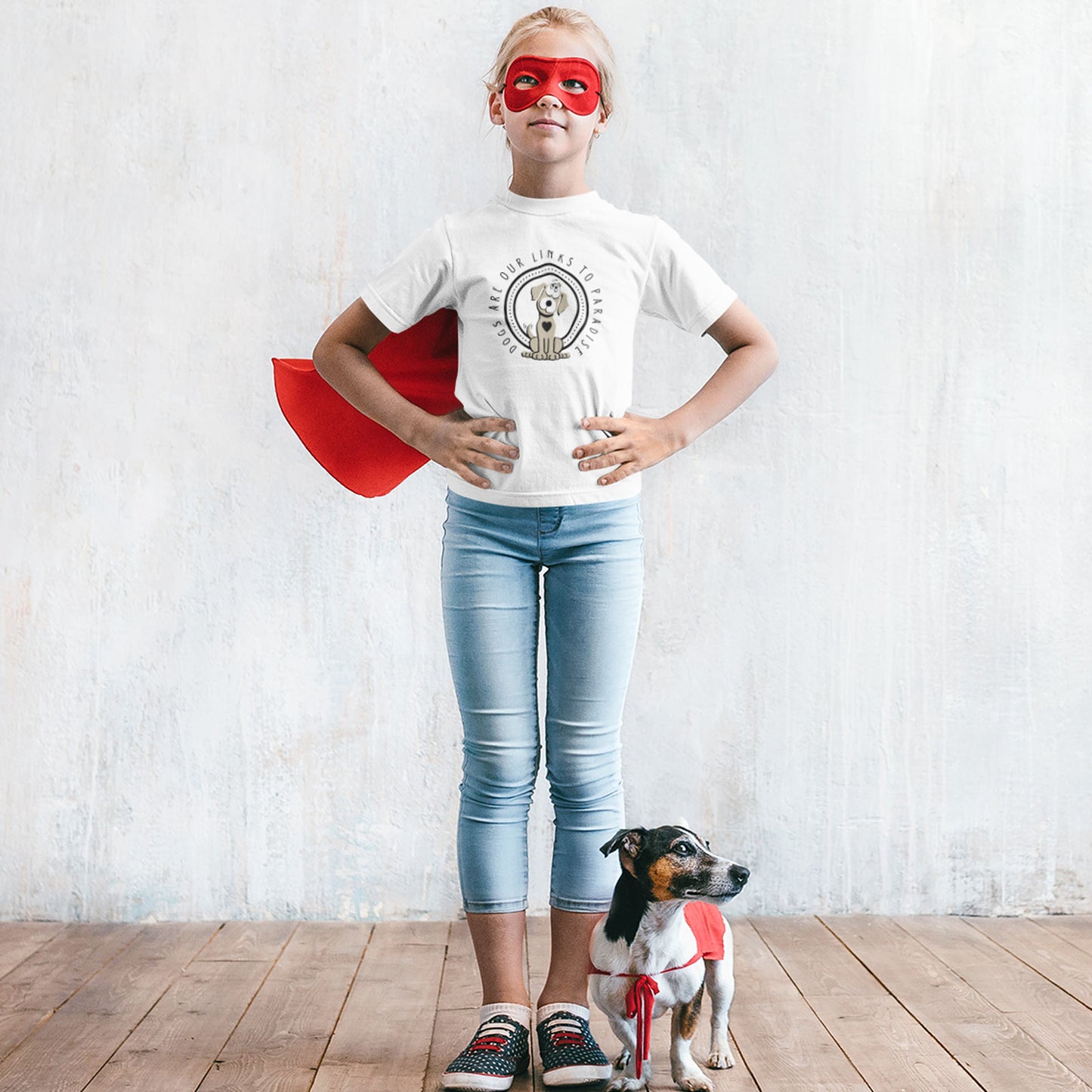 A young supergirl wears a mask, cape and Dogs Pure Love white tee, showcasing the print 'Paradise Is,' as she stands next to her Jack Russel Terrier, against a white background.