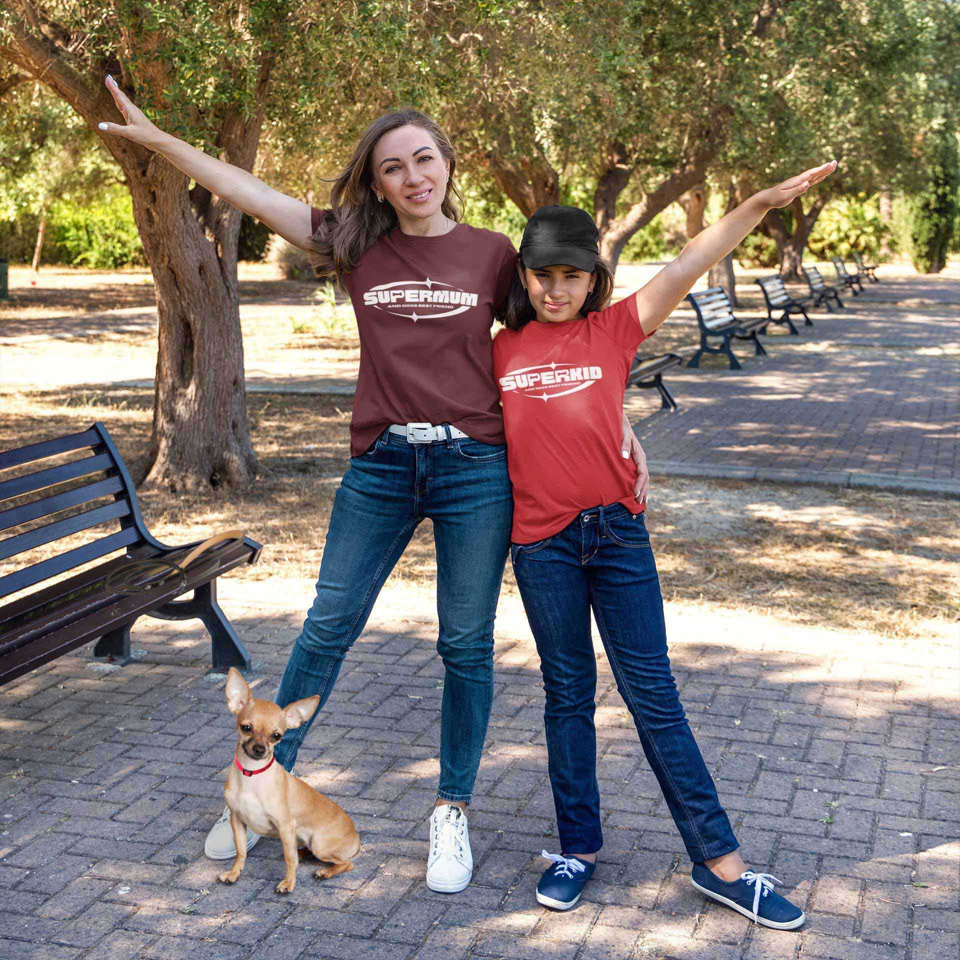  A mother and daughter stand side by side on a boulevard pathway, both sporting their Dogs Pure Love 'Supermum and Superkid' tees, with their arms raised in the air, accompanied by their Chihuahua.