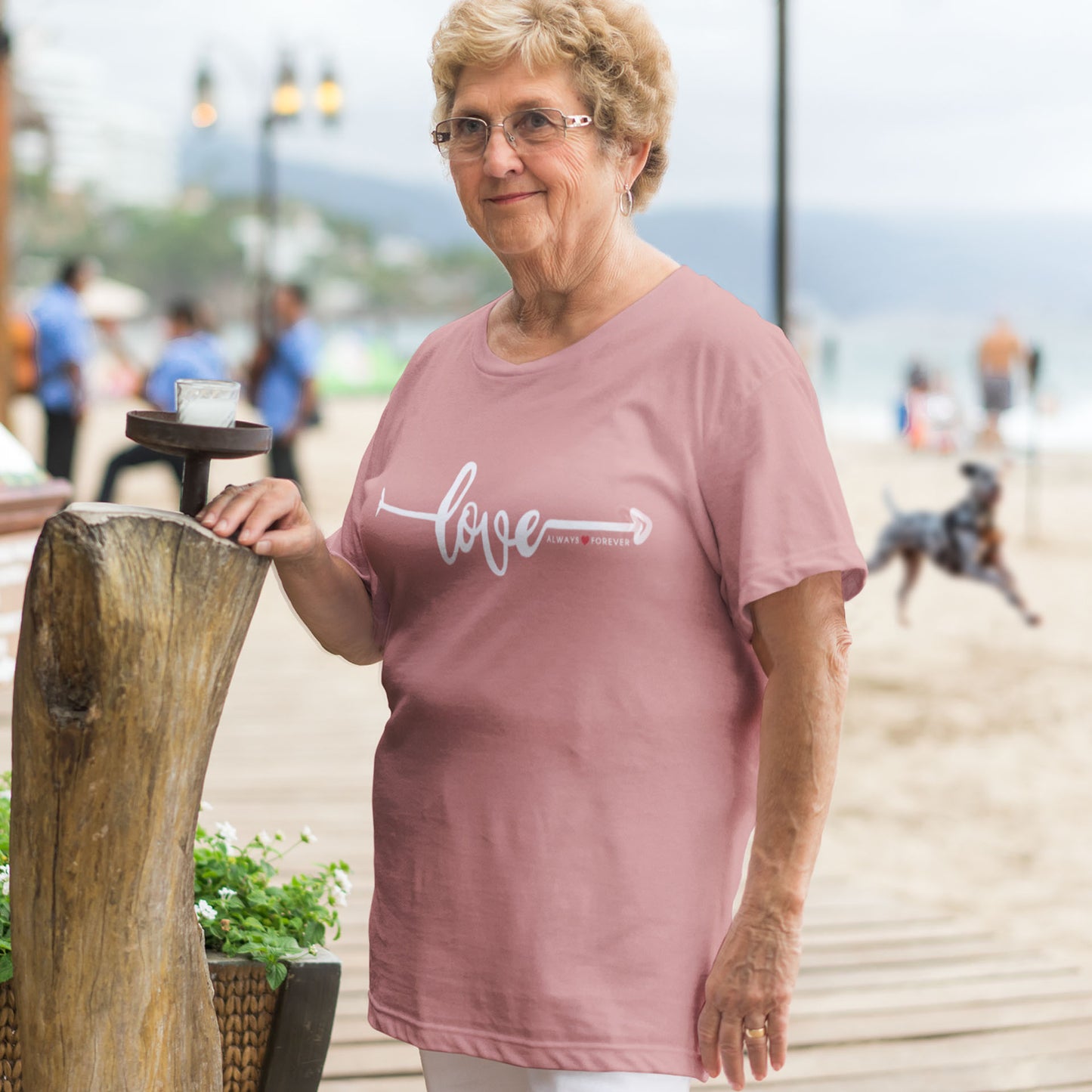  A woman wearing a heather mauve Dogs Pure Love tee with the word "Love" and an arrow stands near a cafe, touching an upright log, while a dog runs on the beach behind her.