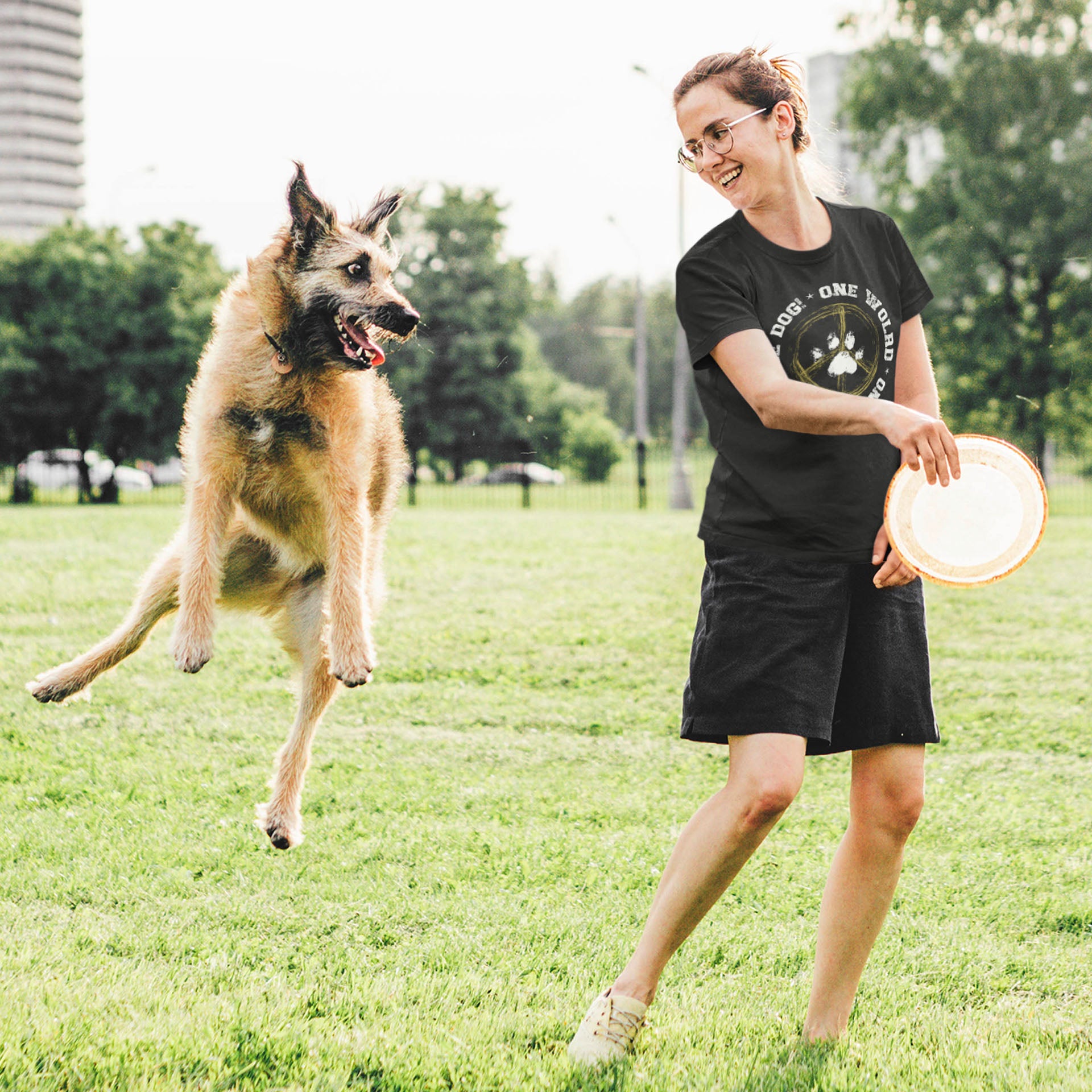 In a park, a woman, dressed in a Dogs Pure Love, 'One World - One Dog' unisex dog t shirt, prepares to throw a frisbee to her dog, who eagerly jumps up to catch it.