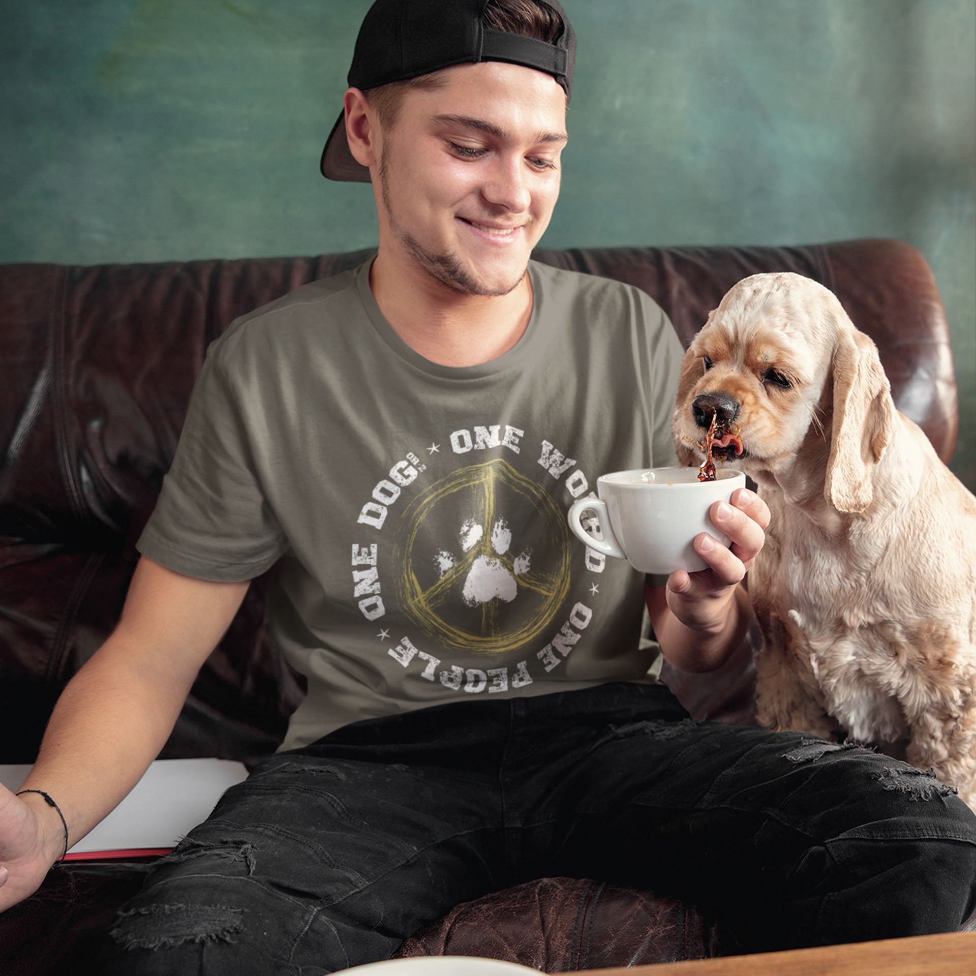 A man, dressed in a Dogs Pure Love, 'One World - One Dog' unisex t shirt, sits on a couch, sharing his coffee cup with his Cocker Spaniel.