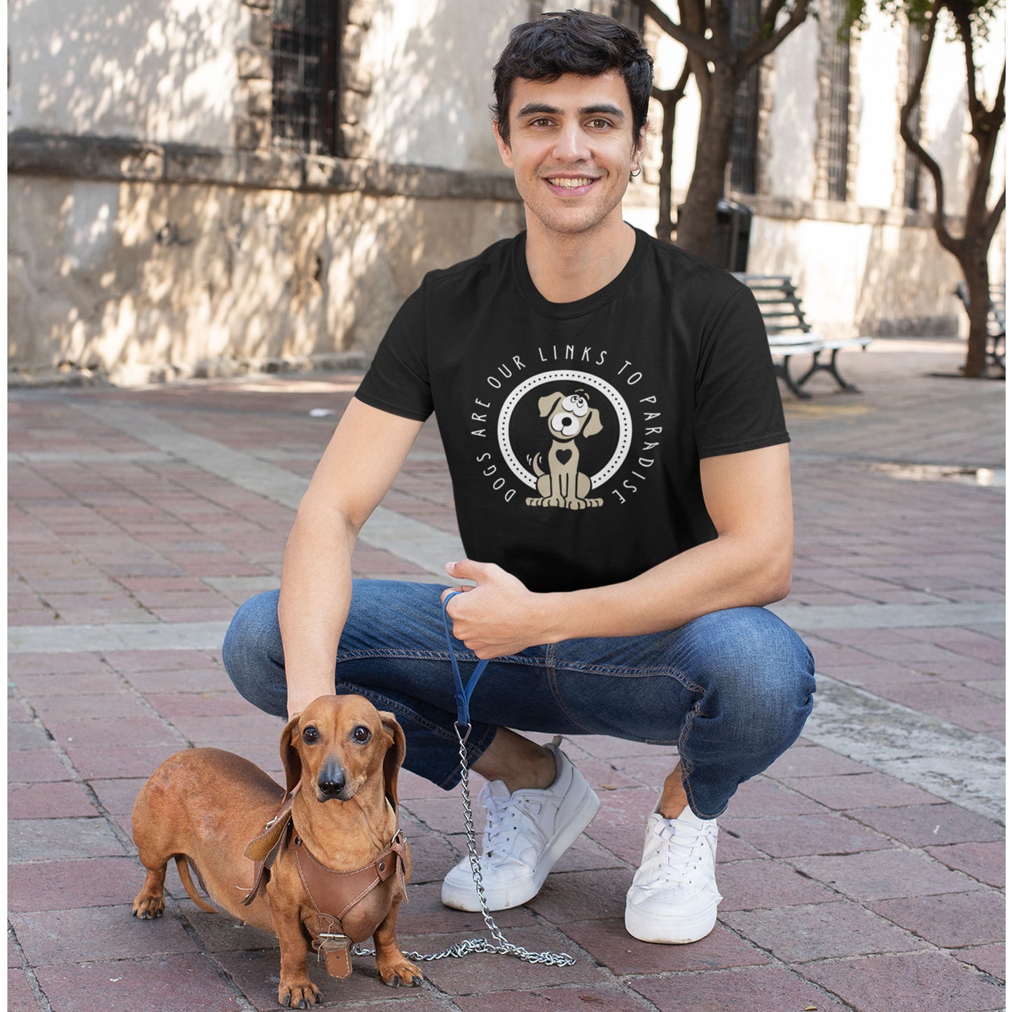  A man, donning a black 'Dogs Pure Love, Dogs are Paradise' unisex tee, crouches down to affectionately pat his Dachshund on a leash while on a tiled footpath.