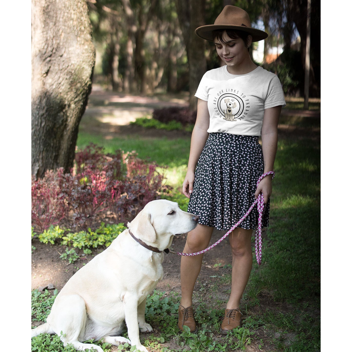  In a park, a woman dressed in a white 'Dogs Pure Love, Dogs are Paradise' unisex tee stands, her gaze directed downwards towards her Labrador Retriever on a leash.