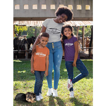 A mother, son and daughter stand together wearing Dogs Pure Love, 'Supermum and Superkid' tees. Their dog lays beside the son, and in the background there are horses in a stable.