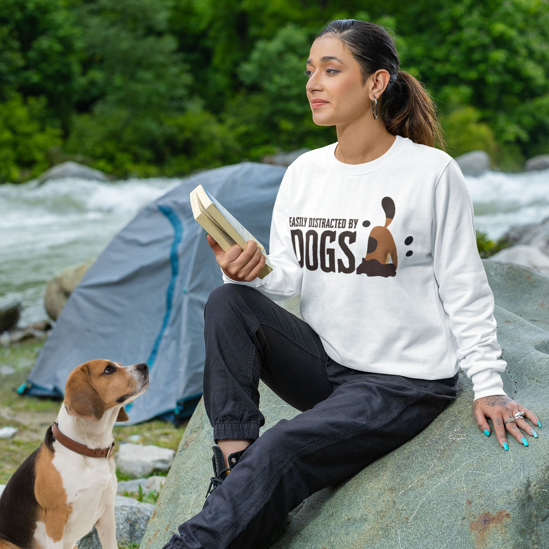 A woman, clad in a 'Dogs Pure Love, Easily Distracted by Dogs' white unisex sweatshirt, perches on a rock with a book in hand, while her Beagle sits admirably in front of her, gazing up. In the background, a tent stands near a tranquil riverbed.