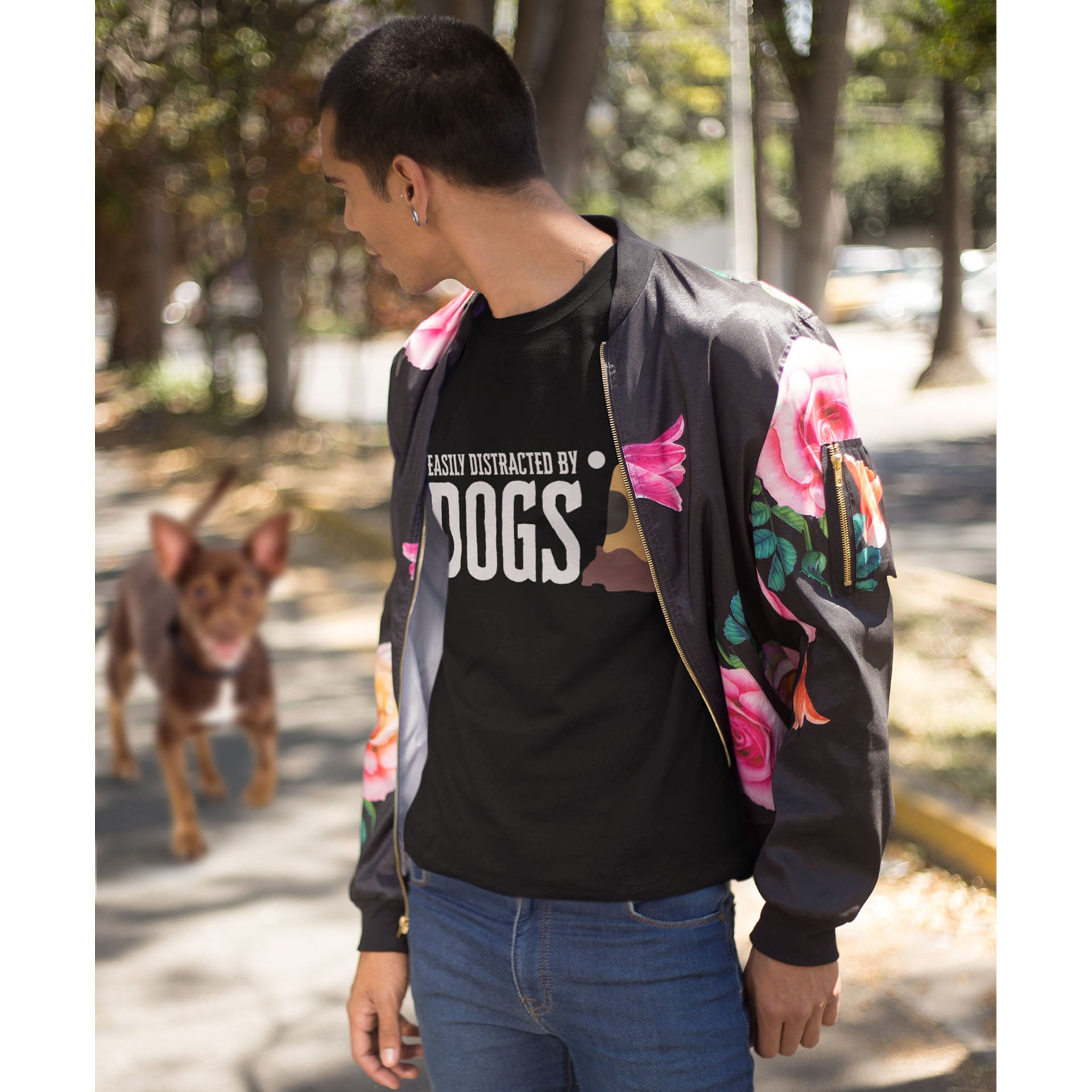 A man, dressed in a unisex black 'Dogs Pure Love, Easily Distracted by Dogs' sweatshirt and a floral jacket, turns to find a dog following him along a pathway.