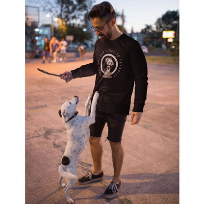 A man stands in an outdoor court area wearing a Dogs Pure Love sweatshirt, holding a stick, while his Jack Russell dog jumps up on him.
