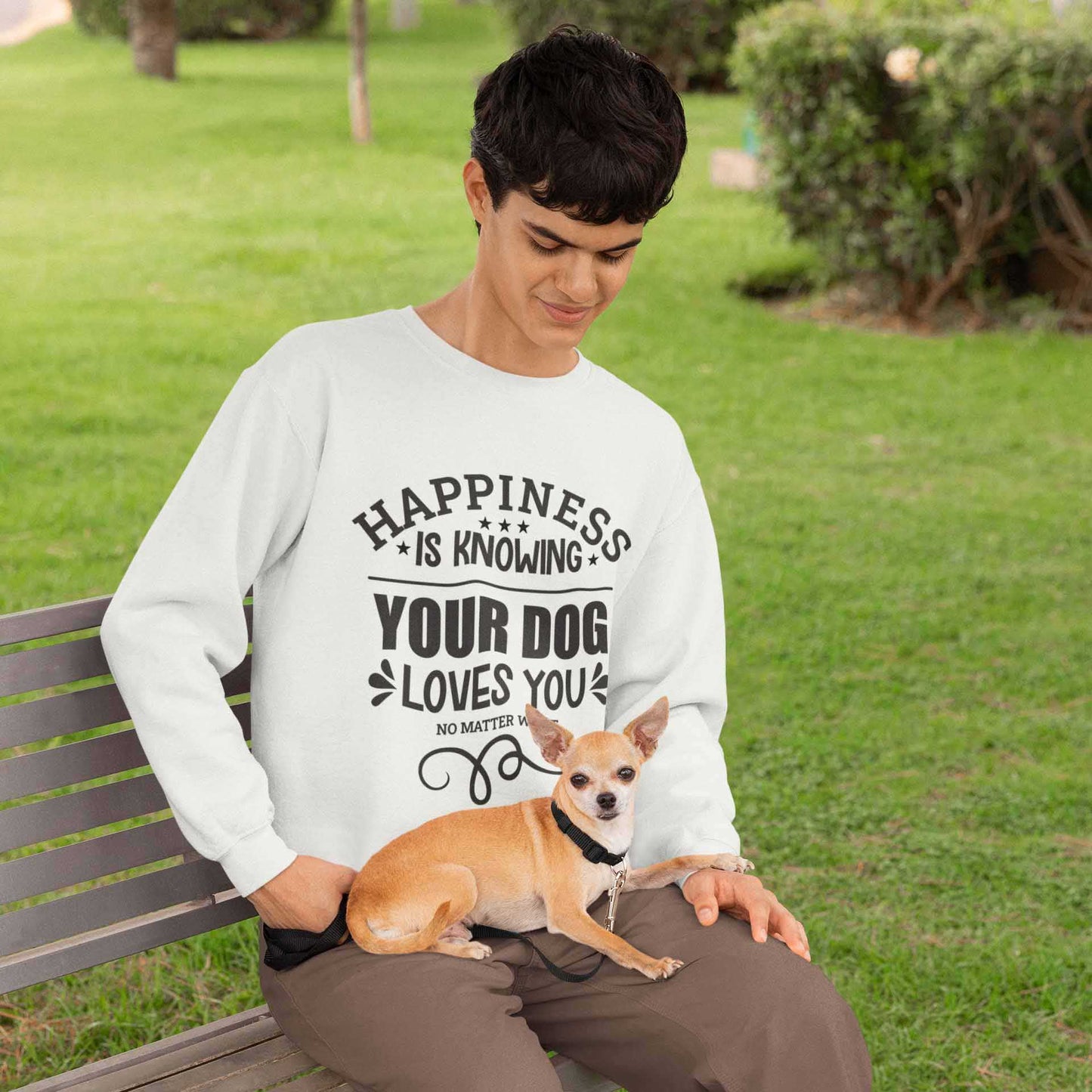 A young man, dressed in a white 'Dogs Pure Love' sweatshirt, sits on a park bench with his Chihuahua nestled on his lap.