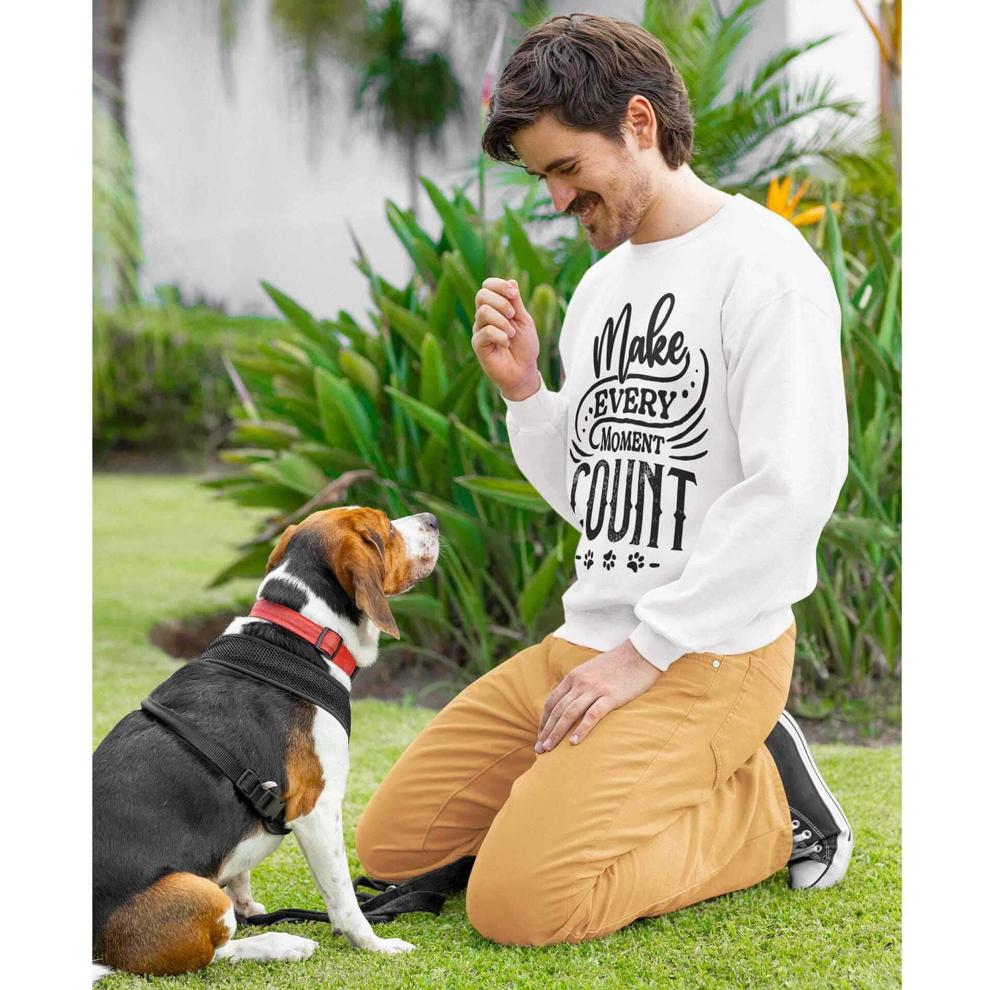  A man wearing a white Dogs Pure Love unisex sweatshirt, adorned with the slogan 'Make Every Moment Count,' kneels on the lawn next to his Beagle, who looks up at him.