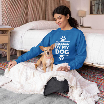 A woman sits on the floor of her bedroom next to her bed wearing a Dogs Pure Love sweatshirt with the text 'Rescued by my Dog,' while her Chihuahua sits on her lap.