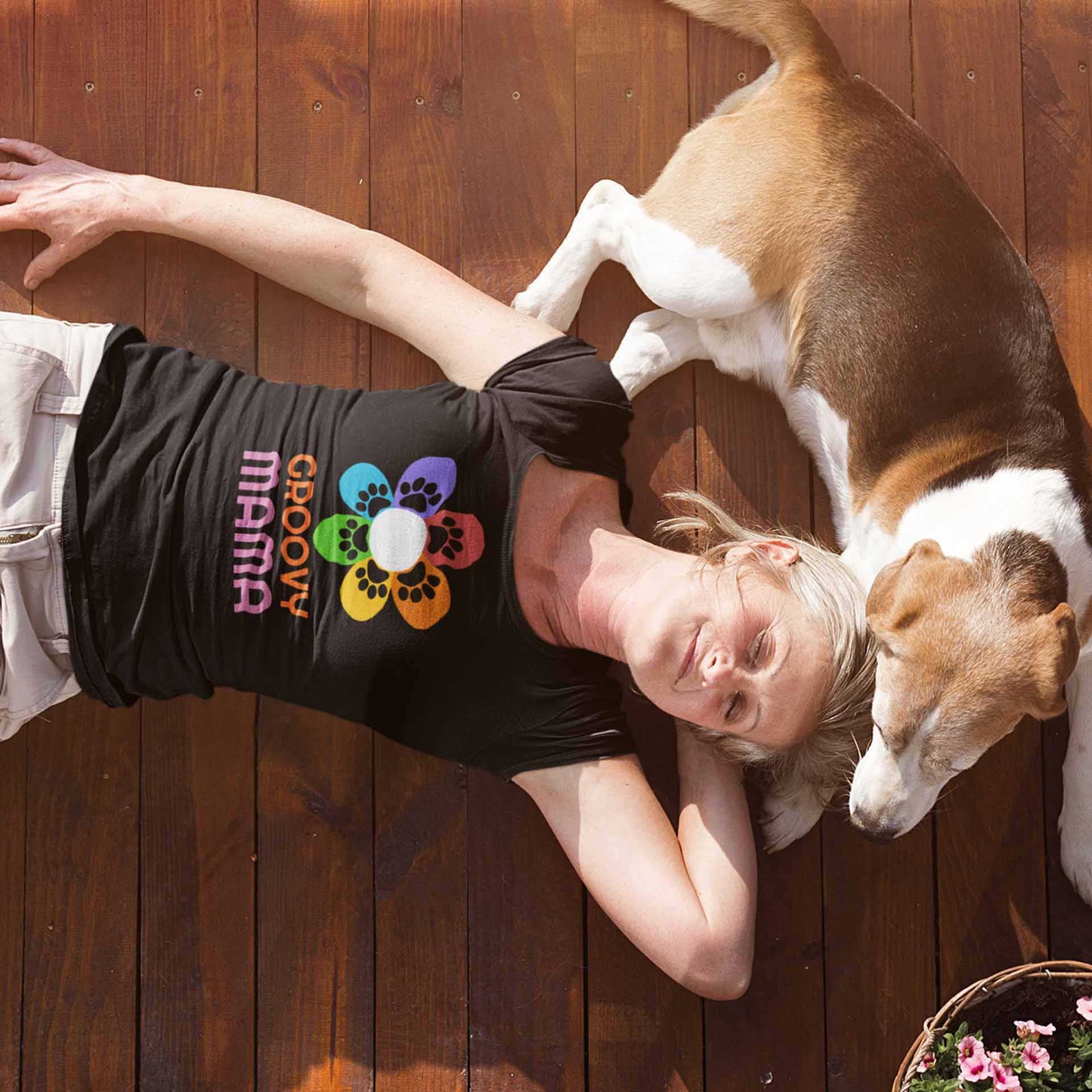 A woman lays on her back on the floor with her dog next to her. She is wearing a Dogs Pure Love black t-shirt that has a colorful flower on it with paw prints and the words 'Groovy Mama' on it. 