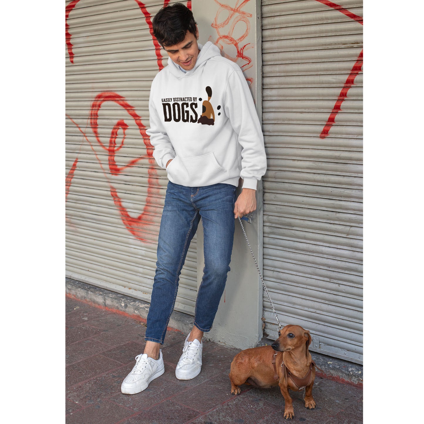 A man, sporting a 'Dogs Pure Love, Dog Dig' hooded sweater, leans against graffiti-covered roller doors, looking down at his Dachshund on a lead.