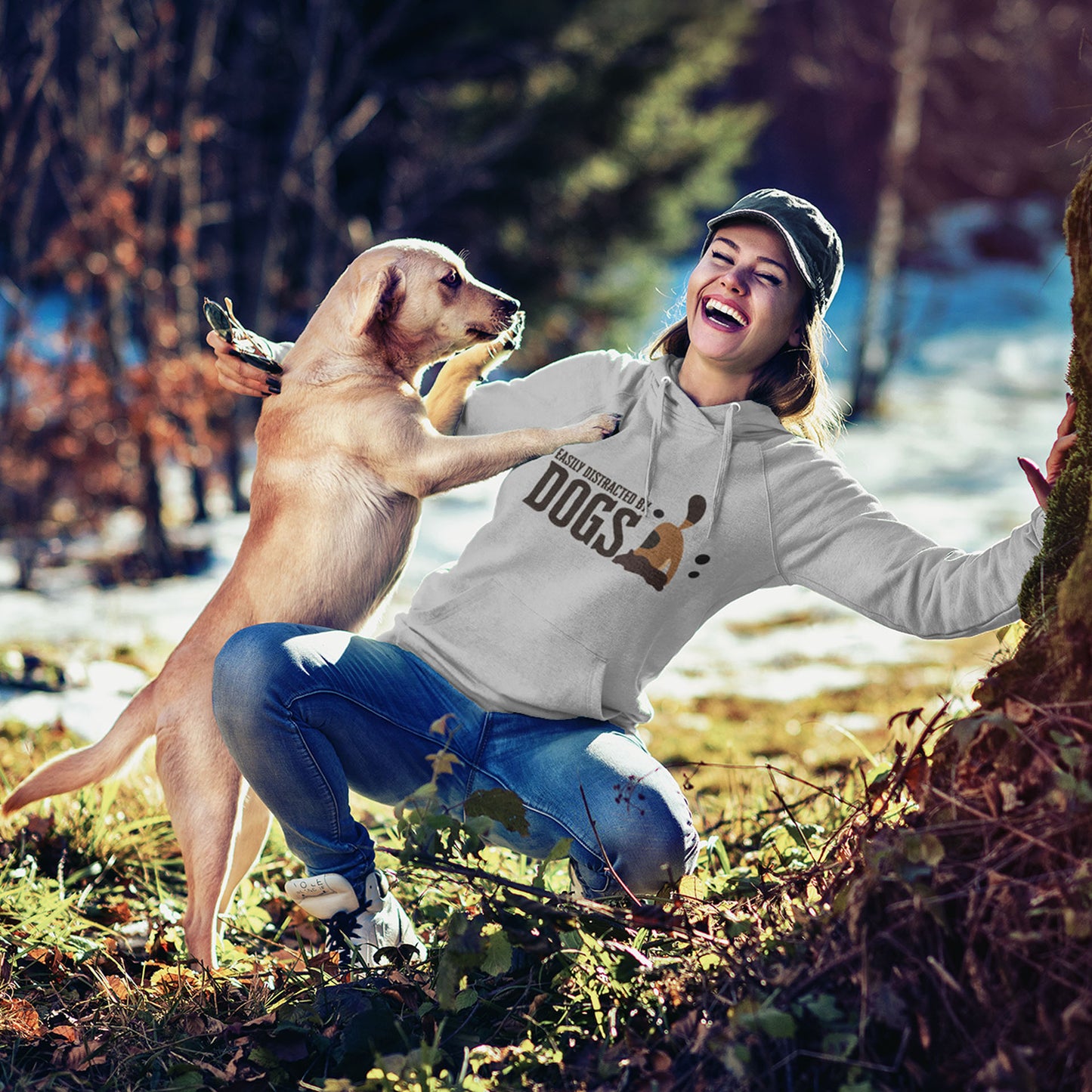 A woman, donning a 'Dogs Pure Love Dog Dig' hooded sweater, crouches down laughing, leaning against a tree, as her Labrador Retriever jumps up on her.