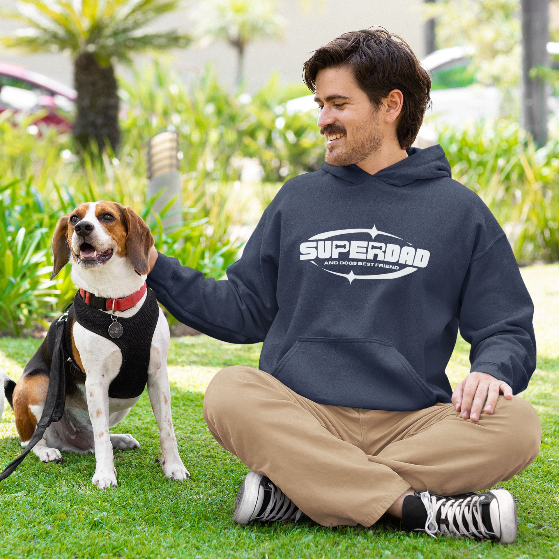  A man sits with his legs crossed next to his Beagle on the grass, he wears a 'Superdad' sweatshirt by Dogs Pure Love.