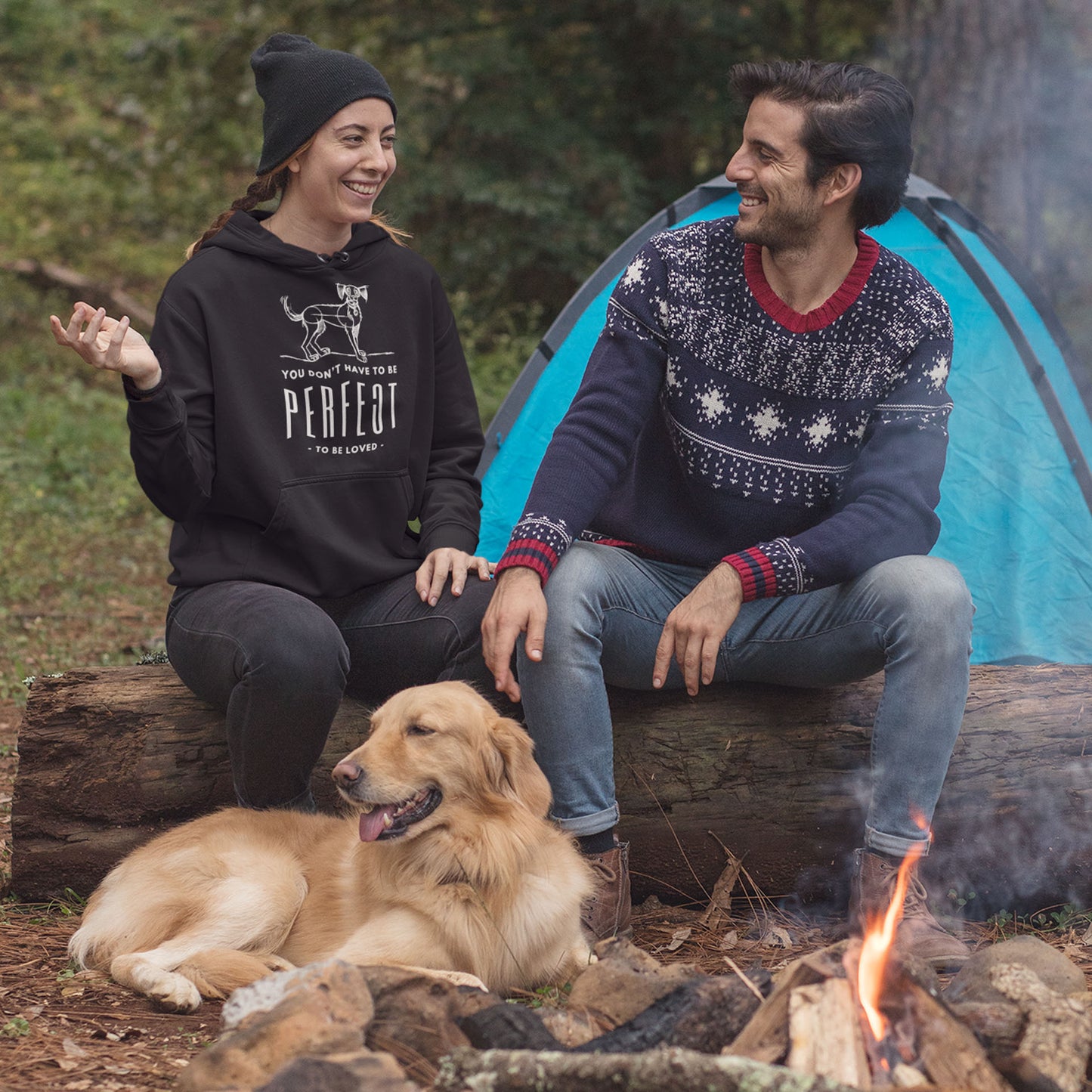 In a scenic forest setting with a tent in the background, a couple sits on a log around a fire with their Golden Retriever. The woman wears a Dogs Pure Love 'Unperfected' hoodie.