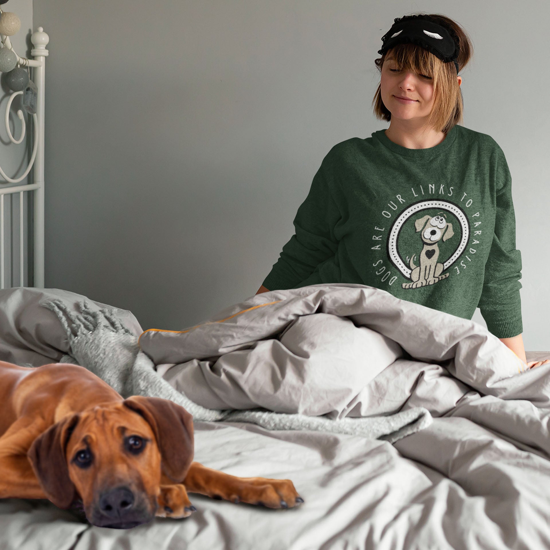 A woman sits upright in a bed, an eyemask resting on her forehead,  she is clad in a Dogs Pure Love sweatshirt featuring the enchanting print 'Dogs are Paradise,' while her loyal dog lays close by.