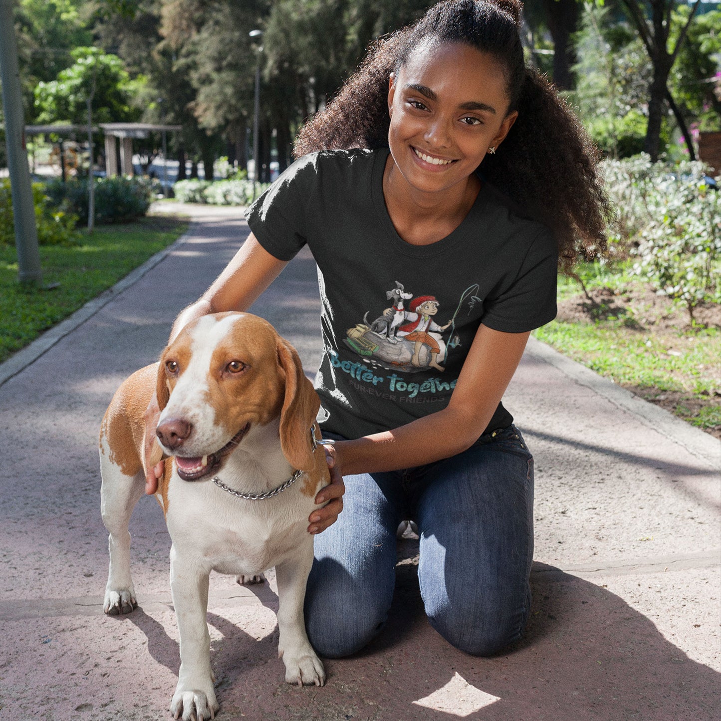 A young woman with curly hair wearing a 'Dogs Pure Love Better Together' unisex tee, kneels on a park footpath holding her Beagle dog. 