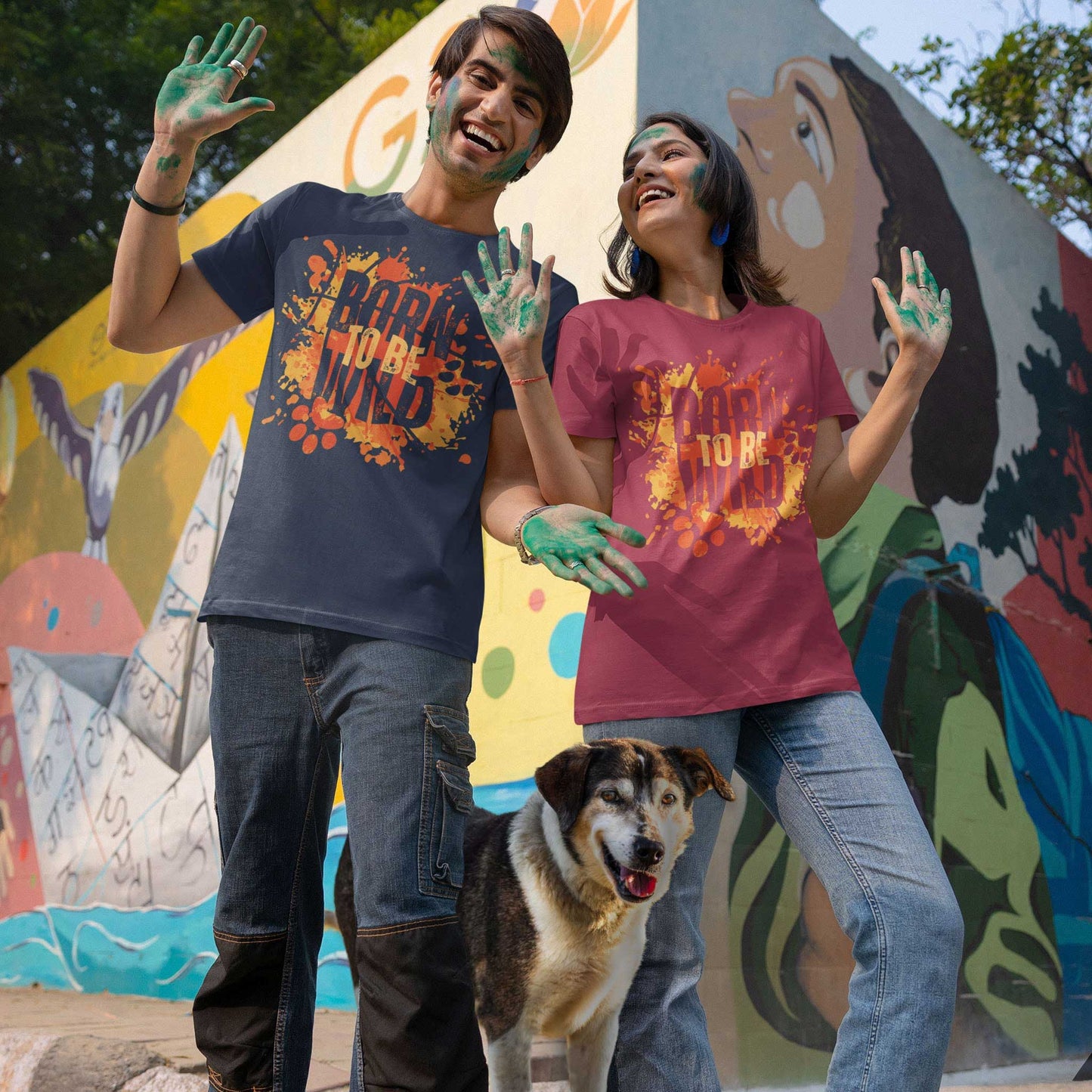 A dog stands between a fun couple covered in paint wearing a 'Dogs Pure Love, Born to be Wild' unisex tee, in-front of a mural wall.