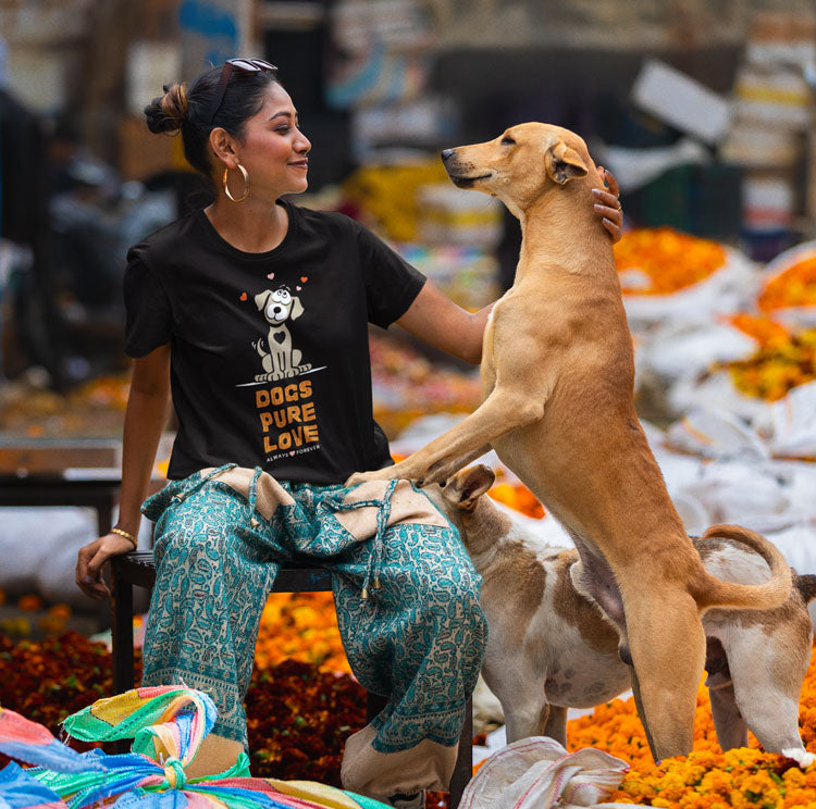  In the bustling market, a woman wearing a 'Dogs Pure Love' unisex tee, sits affectionately patting a dog standing up against her, as another dog stands by.