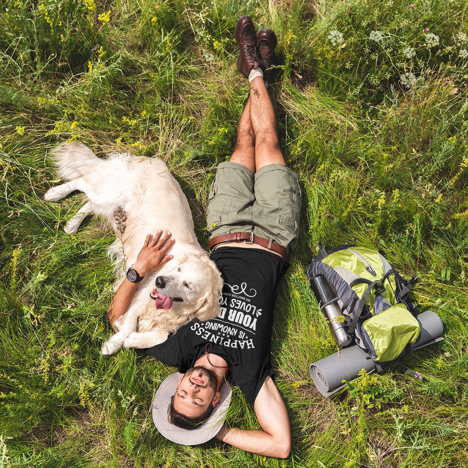  A man wearing a Dogs Pure Love tee lies on the grass next to his backpack, with his legs crossed and one hand behind his head, while the other arm is wrapped around his Golden Retriever.