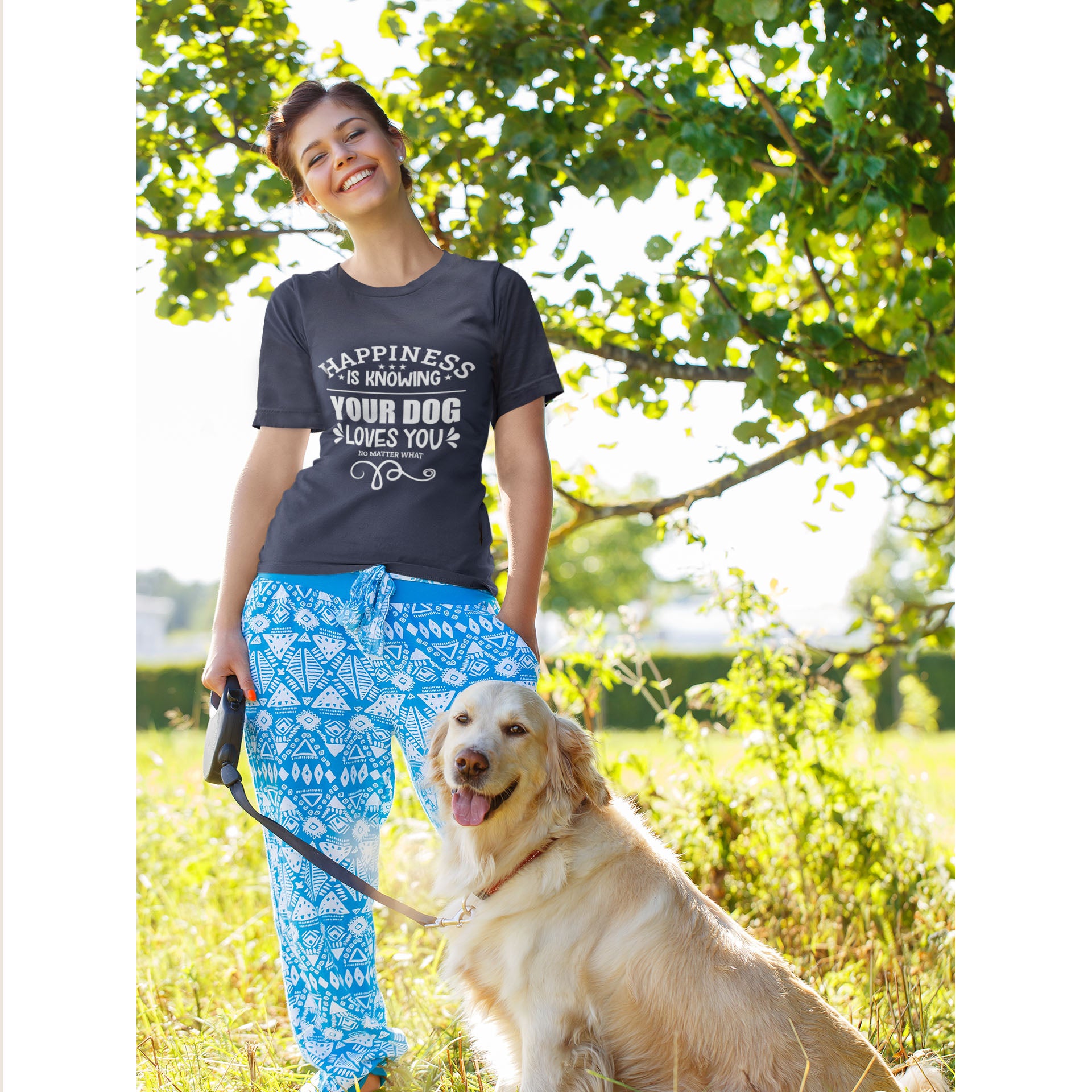  A woman, clad in a Dogs Pure Love navy tee with the slogan 'Happiness is knowing your dog loves you no matter what,' stands in front of a tree, holding the leash of her Golden Retriever.