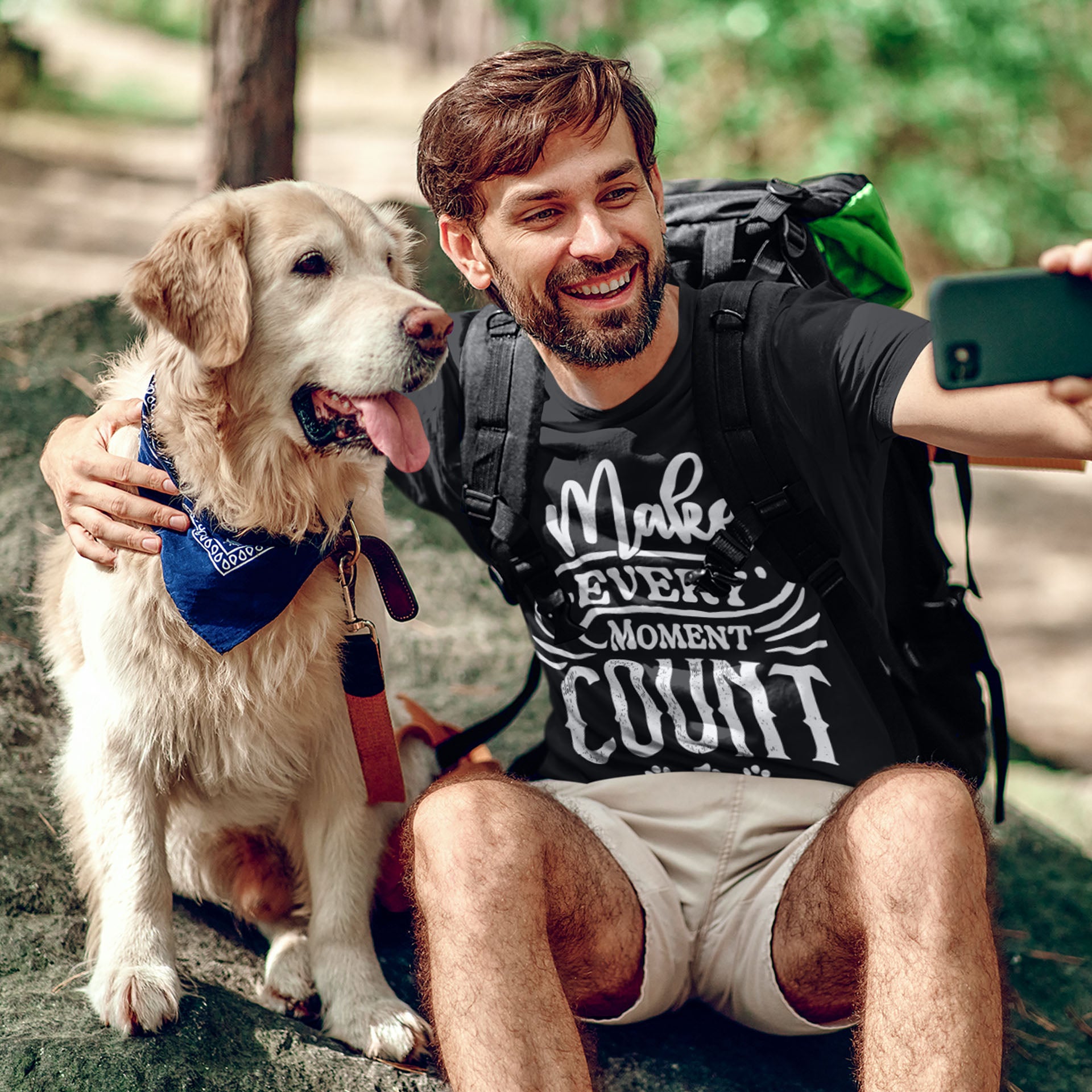 A man wearing a Dogs Pure Love tee with the slogan "Make Every Moment Count" takes a selfie with his Golden Retriever as they sit on a rock in a forest setting.