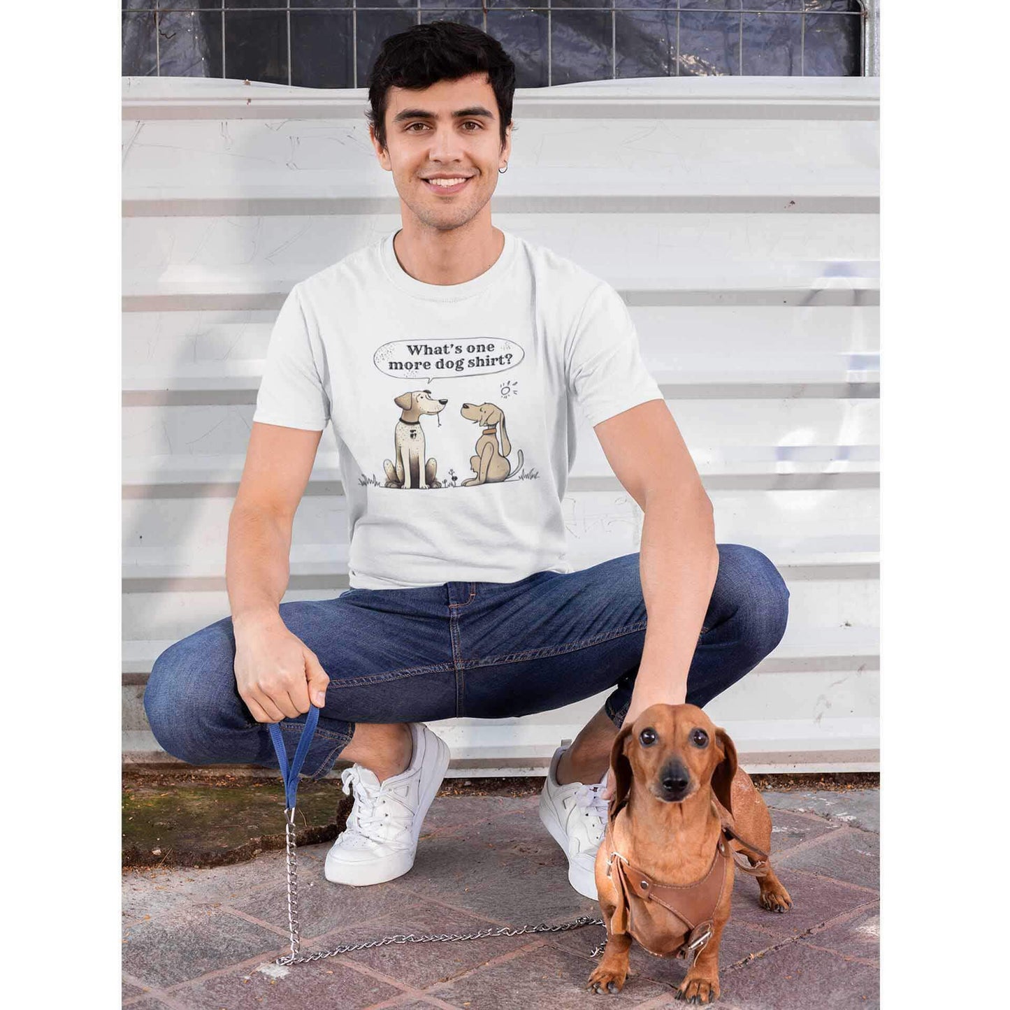 A young man crouches down, his Dachshund stands in front of him.  The man is wearing a white tee with a print of two dogs sitting looking at each other, and one dog has a speech bubble above with the words "Whats one more dog tee."